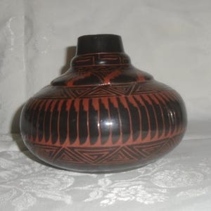 May include: A round, black ceramic vase with a narrow neck and a bulbous body. The vase is decorated with a repeating pattern of red-brown geometric shapes, including triangles and vertical lines. The vase sits on a white surface.