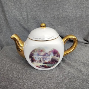 May include: A white ceramic teapot featuring a gold-colored handle, spout, and lid knob. The lid is inscribed with the text "Home Is Where The Heart Is". The body of the teapot displays a colorful illustration of a house surrounded by trees and greenery.
