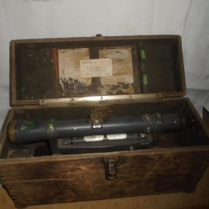 May include: An open, aged wooden box containing a black and gray surveying instrument. The instrument has a cylindrical body with gold and green accents. The box is weathered, with a metal clasp and a label inside the lid.