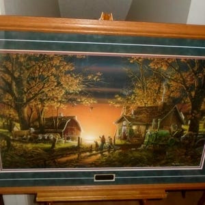 May include: Framed landscape art print depicting a rural scene at sunset. The artwork features a barn, a house, and figures near a body of water, all set against a backdrop of trees and an orange sky. The frame is made of wood.