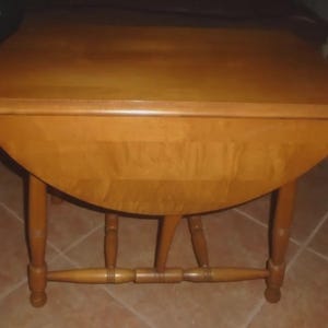 May include: A medium-brown wooden drop-leaf table with a rectangular top and two rounded side leaves. The table features four turned legs connected by a horizontal stretcher base, showcasing a smooth, polished wood grain finish throughout.