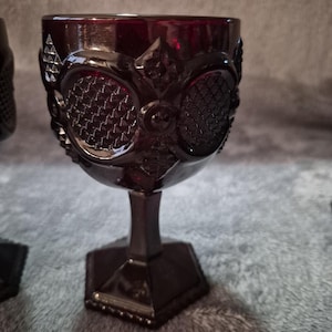 May include: A deep red glass goblet featuring a prominent embossed geometric pattern with circular and diamond-shaped textures. The vessel has a thick stem and a hexagonal base, showcasing a rich, dark crimson hue throughout the entire structure.