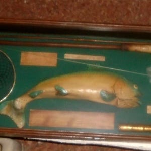 May include: A rectangular wooden display case with a dark green interior containing a carved wooden fish, a fishing net with a wooden handle, a fishing rod, a reel, and several small rectangular wooden plaques with text labels.