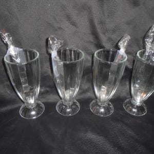 May include: Four clear glass soda fountain glasses with vertical ribbed detailing and a pedestal base. Each glass features a small, clear plastic-wrapped spoon tucked inside the rim, standing upright against the interior wall of the glass.