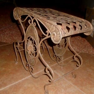 May include: A metal stool with a lattice-patterned top and curved legs. The frame features scrollwork details and oval side panels with a central floral motif. The metal has a weathered, brownish-bronze finish with visible texture throughout.