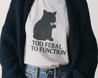May include: A gray crew neck t-shirt featuring a black cat graphic licking its paw. The text "TOO FERAL TO FUNCTION" is printed below the cat. The person is wearing a navy blue cardigan and blue jeans.
