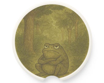 May include: A round car coaster featuring a grumpy-looking frog with crossed arms, set against a forest backdrop. The coaster has a white border and a small notch. The frog and forest are rendered in shades of green and brown.