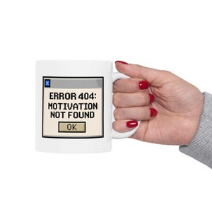 May include: White ceramic mug with a pixelated "ERROR 404: MOTIVATION NOT FOUND" design. The design includes a simulated computer error message with an "OK" button. The mug is held by a person with red nail polish.