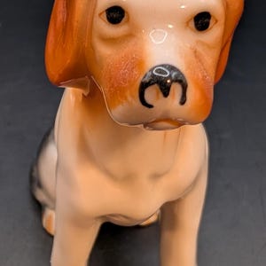 May include: A ceramic figurine of a sitting dog with a smooth, glossy finish. The dog features a light tan body, darker brown ears, and black painted eyes and nose. The canine is depicted in a seated position, facing forward with a neutral expression.