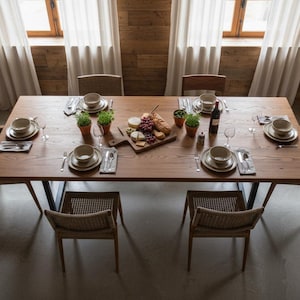 Pode incluir: Uma mesa de jantar de madeira, montada para uma refeição, com cadeiras de vime e talheres para seis pessoas. A mesa é adornada com uma tábua de charcutaria, plantas em vasos e uma garrafa de vinho. A sala tem painéis de madeira e janelas grandes.