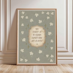 May include: A rectangular wall art print in a wooden frame featuring a sage green background with scattered cream-colored ivy leaves. A central decorative cream-colored plaque contains the text: The secret is to bloom where you are planted.