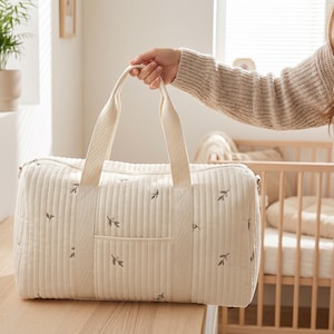 Könnte beinhalten: Cremefarbene, gesteppte Babytasche mit Blättermuster. Die Tasche hat eine rechteckige Form mit einer Vordertasche und zwei Griffen. Die Tasche wird von einer Person in einem beigen Pullover gehalten. Im Hintergrund ist ein Babybett zu sehen.