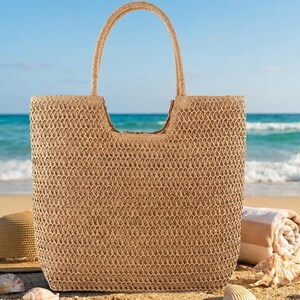 May include: A woven beige tote bag with a rectangular shape and a curved handle. A straw hat, rolled towel, and seashells are on the sand in front of the ocean. The bag is perfect for carrying beach essentials.