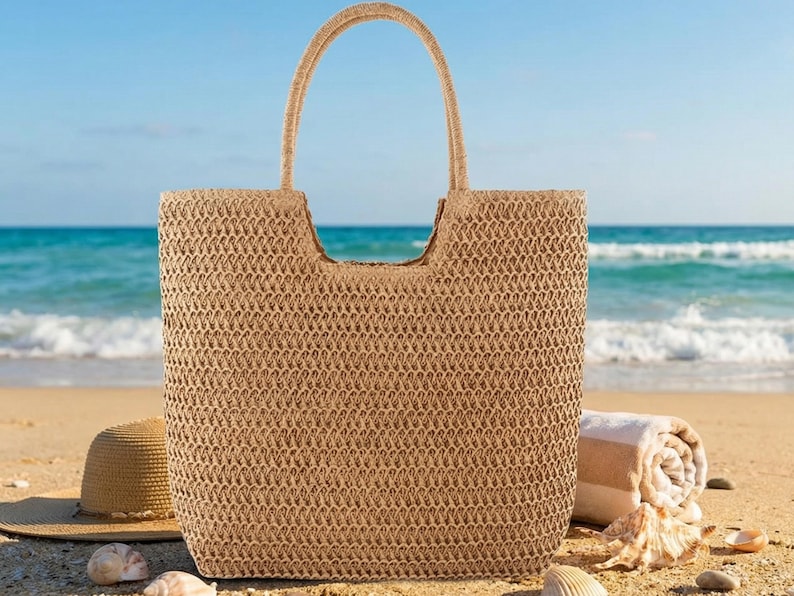 May include: A woven beige tote bag with a rectangular shape and a curved handle. A straw hat, rolled towel, and seashells are on the sand in front of the ocean. The bag is perfect for carrying beach essentials.