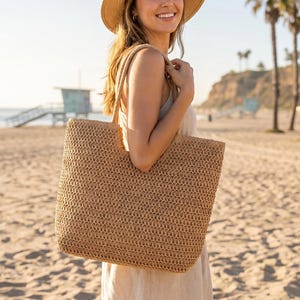 May include: A woven straw tote bag with a rectangular shape and open top. The bag is a light tan color and has a textured appearance. The bag is being carried on a sunny beach day.