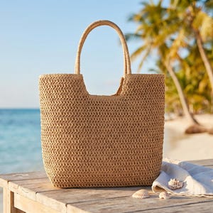 May include: A woven straw tote bag with a rectangular shape and two handles. The bag is light brown and sits on a wooden surface with a white cloth and seashells. The background features a beach and palm trees.