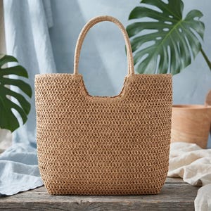 May include: A woven straw tote bag with a rectangular shape and a rounded handle. The bag is a light brown color and sits on a wooden surface. The background includes green leaves and neutral-toned fabrics.