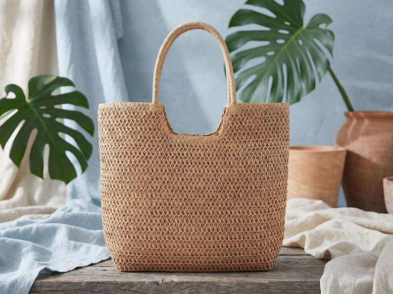 May include: A woven straw tote bag with a rectangular shape and a rounded handle. The bag is a light brown color and sits on a wooden surface. The background includes green leaves and neutral-toned fabrics.
