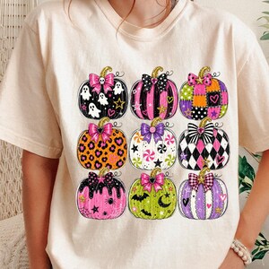 May include: A cream-colored short-sleeve t-shirt featuring a graphic print of nine stylized pumpkins arranged in a grid. Each pumpkin displays a unique pattern, including leopard print, plaid, and polka dots, accented with colorful bows and stars.