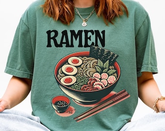 May include: Green t-shirt with the word "RAMEN" in black block letters. The shirt features a graphic of a bowl of ramen with noodles, eggs, seaweed, and other ingredients. Chopsticks are placed next to the bowl.