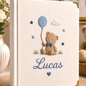 May include: A cream-coloured photo album with gold-tone metal corner protectors. The cover features an embroidered brown teddy bear wearing a blue bow tie, holding a blue balloon, surrounded by blue stars, a white cloud, and the name Lucas in blue.