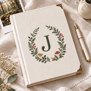 Op de afbeelding: Een crèmekleurig notitieboek met harde kaft en een geborduurde krans van groene bladeren, rode bessen, paddenstoelen en een kleine slak rondom de letter J. Het boek is voorzien van decoratieve goudkleurige metalen hoeken op de kaft.