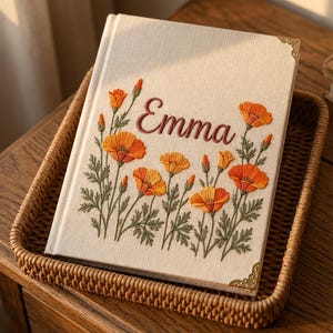 May include: A cream-colored fabric-covered journal featuring an embroidered design of orange poppy flowers and green stems. The name Emma is embroidered in dark red script in the center. The book has gold-toned metal corner protectors.
