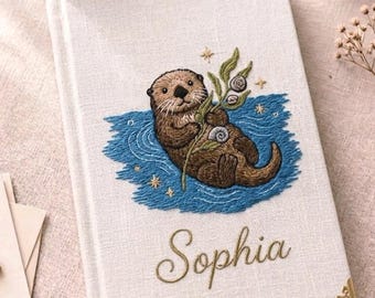 May include: A white fabric-covered journal featuring an embroidered brown otter floating in blue water while holding green seaweed and shells. The name Sophia is embroidered in gold script below the otter. The journal has gold metal corner protectors.