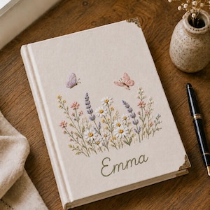 Pode incluir: Um diário revestido de tecido na cor creme com flores silvestres, lavanda e duas borboletas bordadas em tons pastel suaves. O nome Emma está bordado com linha verde na parte inferior e a capa inclui cantos decorativos de metal dourado.