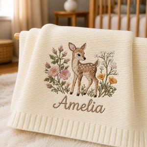 Könnte beinhalten: Eine cremefarbene Strickdecke mit einer Stickerei eines Rehkitzes inmitten von rosa und gelben Wildblumen. Der Name Amelia ist in brauner Schreibschrift unter dem Rehkitz eingestickt. Die Decke hat einen gerippten Randabschluss.