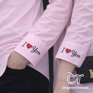 May include: Two pink button-down shirts with the text "I ❤️ You" embroidered on the cuffs. The heart is red, and the text is in black script. The shirts are a light pink color.