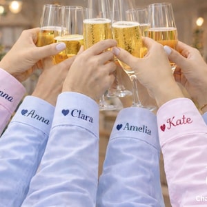 May include: A group of people toasting with champagne flutes. The sleeves of the shirts are visible, embroidered with names like "Anna", "Clara", "Amelia", and "Kate". The shirts are in shades of pink and blue.