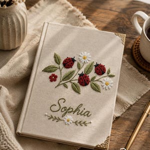 Puede incluir: Un diario con cubierta de tela beige que presenta mariquitas rojas, hojas verdes y margaritas blancas bordadas. El nombre Sophia está bordado en cursiva verde debajo del diseño floral. El libro tiene protectores de esquina de metal dorado.