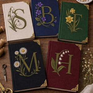 May include: Five rectangular hardcover journals featuring embroidered floral designs and large letters. The covers are cream, navy, dark green, black, and burgundy, each with gold-toned metal corner protectors and unique botanical motifs like daisies and lilies.