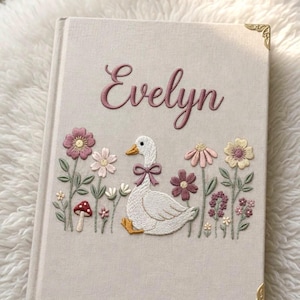 以下が含まれることがあります： ピンク色の筆記体で「Evelyn」と刺繍されたベージュ色の本の表紙。ピンクのリボンをつけた白いガチョウと、ピンク、紫、黄色の刺繍の花が描かれています。金色のコーナーアクセントがエレガントさを添えています。