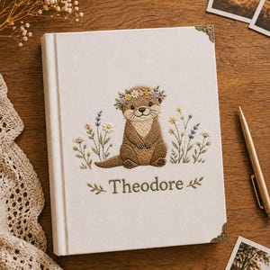 Pode incluir: Um diário revestido de tecido branco com uma lontra castanha bordada a usar uma coroa de flores, rodeada de flores silvestres bordadas. O nome Theodore está bordado a verde por baixo da lontra. O diário tem cantos metálicos decorativos.