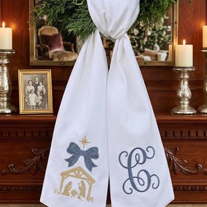 Puede incluir: Bufanda navideña blanca con lazo bordado azul y escena del nacimiento. El otro lado tiene una letra "C" bordada en azul. La bufanda está drapeada sobre una corona verde, con velas y una foto enmarcada en el fondo.
