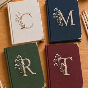 Puede incluir: Cuatro diarios rectangulares con cubiertas de tela en color crema, azul marino, verde bosque y burdeos. Cada diario presenta una letra mayúscula bordada C, M, R o T acentuada con ramitas de flores blancas y esquineros de metal dorado.