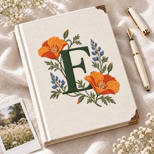 Op de afbeelding: Een crèmekleurig notitieboek met harde kaft, voorzien van een geborduurde donkergroene letter E omringd door oranje klaprozen en blauwe lupinebloemen. Het notitieboek heeft goudkleurige metalen hoekbeschermers en een crèmekleurige vulpen.