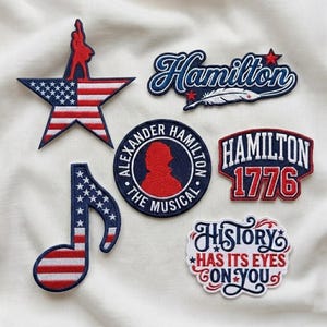 Op de afbeelding: Collectie geborduurde patches met patriottische thema's. Ontwerpen omvatten een ster met de Amerikaanse vlag, een muzieknoot met sterren en strepen, en patches met het woord "Hamilton" en de zin "History Has Its Eyes On You."