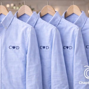 May include: Four light blue button-down shirts hanging on wooden hangers. Each shirt has the embroidered initials "C♥D" on the left chest. The shirts are displayed against a blurred background with Christmas lights and the ChaptersThreads logo.