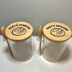 May include: Two clear glass jars with wooden lids and small wooden spoons. The lids are engraved with the words "BART'S STARTER" and "BART'S DISCARD" in black. The jars are designed for food storage.