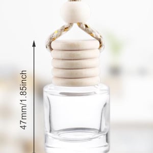 Op de afbeelding: Een heldere glazen auto-luchtverfrisserfles met een houten dop en een ophangtouw. De fles is 4,7 cm hoog en 2,6 cm breed. Het touw is beige en goudkleurig.