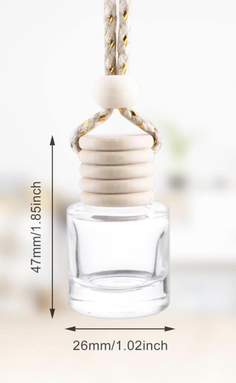 Op de afbeelding: Een heldere glazen auto-luchtverfrisserfles met een houten dop en een ophangtouw. De fles is 4,7 cm hoog en 2,6 cm breed. Het touw is beige en goudkleurig.