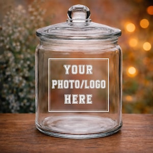 Peut inclure: Un bocal en verre transparent avec un couvercle et un bouton sur le dessus. Le bocal a un cadre rectangulaire blanc avec le texte "YOUR PHOTO/LOGO HERE" au centre. Le bocal est posé sur une surface en bois.