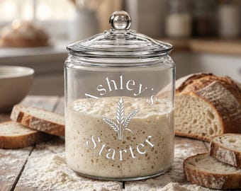 May include: A clear glass jar with a lid containing sourdough starter. The front of the jar features white text that reads Ashley's Starter with a graphic of three wheat stalks in the center. The jar is filled with a bubbly beige mixture.