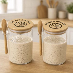 May include: Two clear glass jars filled with beige sourdough starter, each featuring a light wood lid with a black graphic of wheat and the text ASHLEY'S STARTER or ASHLEY'S DISCARD. Each jar includes a small wooden spoon held by a clear side clip.