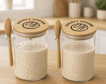 May include: Two clear glass jars filled with beige sourdough starter, each featuring a light wood lid with a black graphic of wheat and the text ASHLEY'S STARTER or ASHLEY'S DISCARD. Each jar includes a small wooden spoon held by a clear side clip.