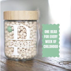 May include: A clear glass jar with a wooden lid filled with small, light-colored wooden beads. The jar has a white monogram with the letter "D" and the name "PARKER". A green scalloped sign reads "ONE BEAD FOR EVERY WEEK OF CHILDHOOD".