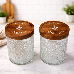 May include: Two clear glass jars with textured sides and wooden lids. The lids are engraved with the words "Erin's Starter" and "Erin's Discard" along with a wheat stalk design. The jars are designed for kitchen use.